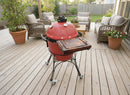 A BBQ Boards® Kamado Joe Classic Joe I Side Cutting Board is attached to a red ceramic grill on wheels on a wooden deck, surrounded by wicker patio furniture, potted plants, and a wooden fence—ideal for BBQ prep.