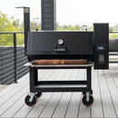 A large black outdoor smoker grill with a thermometer, digital controls, and pellet smoker accessories sits on a wooden patio. Red-rimmed wheels and a BBQ Boards® Lone Star Grillz 20" x 42" Front Cutting Board shelf add function, with trees beyond.
