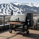 A black outdoor grill with a wooden prep area features a BBQ Boards® Lone Star Grillz 20" x 42" Front Cutting Board on the deck, with snowy mountains and a ski resort visible beneath a partly cloudy sky.
