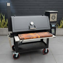 A large black outdoor smoker grill features a BBQ Boards® Lone Star Grillz 20" x 42" Front Cutting Board on the front tray, a digital control panel, and a "7 Star BBQ" logo, set on a patio with plants and a dark fence behind.