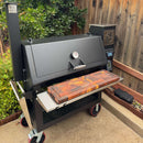 A large black smoker grill with a built-in thermometer and digital control panel sits on the patio by a wooden fence, featuring a vibrant BBQ Boards® Lone Star Grillz 20" x 42" Front Cutting Board on its front shelf.