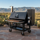 A BBQ Boards® Lone Star Grillz 20" x 42" front cutting board is featured on a large black outdoor smoker grill with a digital control panel, set on a stone patio amid desert plants and mountain views.