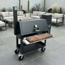 A modern black outdoor smoker grill on wheels features the BBQ Boards® Lone Star Grillz 20" x 42" Front Cutting Board by BBQ Boards® on its side shelf, placed on a gray-tiled patio with beige sofas and assorted cacti and plants nearby.
