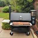 A spacious outdoor smoker grill with a BBQ Boards® Lone Star Grillz 20" x 42" Front Cutting Board, shelves, thermometer, and digital controls stands on a fenced patio surrounded by green grass and shrubs.