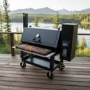A black outdoor smoker grill with a BBQ Boards® Lone Star Grillz 20" x 42" Front Cutting Board rests on a wooden deck, overlooking a scenic lake and forested mountains under a clear sky.