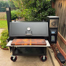 A large outdoor smoker grill with a thermometer, digital control panel, and a BBQ Boards® Lone Star Grillz 20" x 42" Front Cutting Board on the front shelf sits on a patio by a wooden fence and green plants.