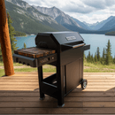 A BBQ Boards® Masterbuilt AutoIgnite Series 545 Side Cutting Board is attached to a black grill on a wooden deck overlooking a lake with mountains and pine trees, ready for prep under a partly cloudy sky.