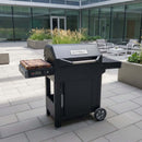 A black Masterbuilt AutoIgnite Series 545 grill stands on a modern patio with gray tile, seating, and tall grasses, featuring a BBQ Boards® Masterbuilt AutoIgnite Series 545 Side Cutting Board for convenient grill-side prep.