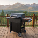A BBQ Boards® Masterbuilt AutoIgnite Series 545 Side Cutting Board sits on a black grill atop a wooden deck, surrounded by pine trees and mountains under a clear sky—ideal for convenient outdoor prep.