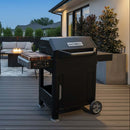 A black Masterbuilt AutoIgnite Series 545 grill stands on a patio deck with the BBQ Boards® Masterbuilt AutoIgnite Series 545 Side Cutting Board, near modern outdoor seating, fire pit, and plants, with a house in the background during daylight.