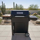 A BBQ Boards® Masterbuilt AutoIgnite Series 545 Side Cutting Board sits attached to a black grill on a tiled patio, with cacti and rocks in the desert landscape under a clear sky.