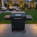 A BBQ Boards® Masterbuilt AutoIgnite Series 545 Side Cutting Board is set up on a patio with outdoor seating, grill-side prep, a fire pit, string lights, and tidy landscaping, creating a cozy backyard ambiance at dusk.