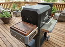 A BBQ Boards® Masterbuilt Gravity Series 800 Side Cutting Board is featured on a black Masterbuilt Gravity Series 800 grill, set on a wooden deck with patio furniture and potted plants for an efficient grill-side workflow.