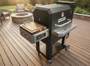 A black Masterbuilt Gravity Series 800 grill features a BBQ Boards® Masterbuilt Gravity Series 800 Side Cutting Board on a wooden deck. Outdoor chairs, a stone fire pit, and green plants line the patio in the background.