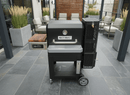 A BBQ Boards® Masterbuilt Gravity Series 800 Side Cutting Board is set up next to a Masterbuilt Gravity Series 800 charcoal grill on a modern patio with gray tile, potted plants, and cushioned seating for efficient outdoor cooking.