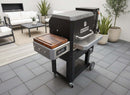 A BBQ Boards® Masterbuilt Gravity Series 800 Side Cutting Board sits on a grill on a gray tile patio, with white cushioned patio furniture and potted plants providing an inviting, efficient setting for outdoor cooking.