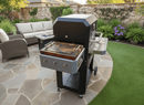 A BBQ Boards® Masterbuilt Gravity Series 800 Side Cutting Board is attached to a modern outdoor grill on a stone patio, surrounded by green plants, artificial turf, and a wicker sofa set in a landscaped backyard.
