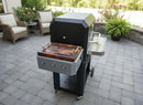 A BBQ Boards® Masterbuilt Gravity Series 800 Side Cutting Board is attached to a black grill on a paved patio. Wicker chairs, a green fern, and flower pots create an inviting backdrop for grilling with BBQ Boards®.