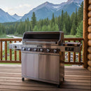 A BBQ Boards® Monument Grills Denali 610 Side Cutting Board by BBQ Boards® is featured on a wooden deck with a scenic mountain view, set beside a stainless steel grill under clear skies.
