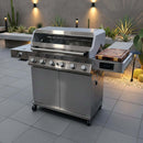 A BBQ Boards® Monument Grills Denali 610 Side Cutting Board is mounted on a large stainless steel grill with five knobs and side burners, displayed on a tiled patio surrounded by cacti and modern wall lights.