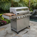 A BBQ Boards® Monument Grills Denali 610 Side Cutting Board is installed on a Monument Grills Denali 610 with six knobs and a canopy lid, placed on a stone patio among lush tropical plants and greenery.