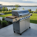 A BBQ Boards® Monument Grills Denali 610 Side Cutting Board sits on a stainless steel grill with six knobs on a patio overlooking a lake and greenery under a partly cloudy sky.