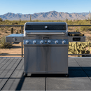 A BBQ Boards® Monument Grills Denali 610 Side Cutting Board sits on a stainless steel gas grill with six knobs on a patio, framed by cacti and mountains in the background beneath a clear blue sky.