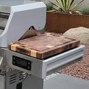 The BBQ Boards® Monument Grills Denali 610 Side Cutting Board by BBQ Boards® sits on the grill’s metal side board, bordered by rocks and plants.