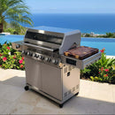 A BBQ Boards® Monument Grills Denali 610 Side Cutting Board from BBQ Boards® is featured on a tiled patio with side shelves, overlooking a pool, tropical plants, and an ocean view beneath a clear blue sky.