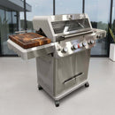 A BBQ Boards® Monument Grills Mesa 405BZ Side Cutting Board sits on a modern stainless steel grill with four knobs, glass lid, and side shelves, positioned on a patio in front of large glass windows.