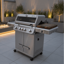 A BBQ Boards® Monument Grills Mesa 405BZ Side Cutting Board is mounted on a modern grill with four control knobs and a thermometer, set on a tiled patio surrounded by ambient lighting and tall plants.