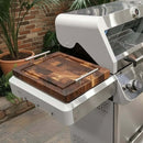 A BBQ Boards® Monument Grills Mesa 405BZ Side Cutting Board sits on the grill’s side shelf, set up on a tiled patio near potted plants and a brick wall.
