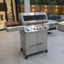 A BBQ Boards® Monument Grills Mesa 405BZ Side Cutting Board is paired with a modern stainless steel grill on the patio, next to a sofa and fire table surrounded by plants and outdoor lighting.