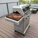 A BBQ Boards® Monument Grills Mesa 405BZ Side Cutting Board is shown attached to a Mesa 405BZ gas grill on a wooden deck, with patio furniture and a glass railing in the background.
