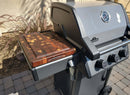 A silver outdoor gas grill with three black knobs, a temperature gauge, and a BBQ Boards® Napoleon Freestyle Side Cutting Board sits on a paved patio beside beige walls and ornamental grasses.