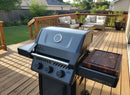 A black gas grill with three knobs and a built-in thermometer stands on a wooden deck, featuring the BBQ Boards® Napoleon Freestyle Side Cutting Board for prep. The deck has patio furniture and overlooks a backyard with a wooden fence.