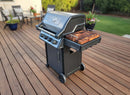A black four-burner outdoor gas grill with a BBQ Boards® Napoleon Freestyle Side Cutting Board by BBQ Boards® is on a wooden deck, surrounded by patio furniture and plants.