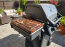 The BBQ Boards® Napoleon Freestyle Side Cutting Board by BBQ Boards® sits on a modern outdoor grill’s side board in a sunny patio, surrounded by potted plants and patio furniture.