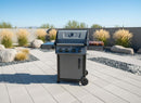 A modern outdoor gas grill with four burners and side shelves features a BBQ Boards® Napoleon Freestyle Side Cutting Board for grill-side prep, set on a tiled patio bordered by rocks, gravel, tall grass, and a concrete wall under a clear blue sky.