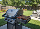 A BBQ Boards® Napoleon Freestyle Side Cutting Board by BBQ Boards® sits next to a three-knob gas grill with a thermometer on a backyard patio, surrounded by a lawn, vibrant flowerbeds, trees, and outdoor wicker furniture.
