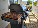 A BBQ Boards® Napoleon Freestyle Side Cutting Board by BBQ Boards® sits on a gas grill with a closed lid on a wooden deck near a white railing, ideal for grill-side prep, surrounded by potted plants and greenery.