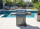 A modern gas grill with blue control knobs and a BBQ Boards® Napoleon Freestyle Side Cutting Board stands on a patio near a pool, surrounded by outdoor seating, potted plants, and palm trees on a sunny day.