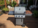 A modern three-burner gas grill with a thermometer and a BBQ Boards® Napoleon Freestyle Side Cutting Board sits on a wooden deck, surrounded by outdoor furniture and potted plants—perfect for easy grill-side prep.