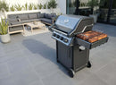 A modern black gas grill with a BBQ Boards® Napoleon Freestyle Side Cutting Board sits on a spacious gray-tiled patio near an outdoor seating area with a gray sofa, chairs, and coffee table, all surrounded by potted plants.