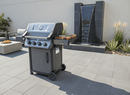 A modern gas grill with a BBQ Boards® Napoleon Freestyle Side Cutting Board stands on a tiled patio near a gray stone water feature and plants. In the background, there’s a gray outdoor sofa and a food-laden table.