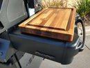 The BBQ Boards® Napoleon Prestige 500 Side Cutting Board by BBQ Boards® rests on the side board, providing generous grill prep space, with greenery and pavement in the background.