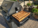 A Napoleon Prestige 500 gas grill with a BBQ Boards® Side Cutting Board rests on a patio with wicker furniture, potted plants, and stone walls in the background.