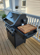 A modern black grill with four knobs and a built-in thermometer stands on a deck. The BBQ Boards® Napoleon Prestige 500 Side Cutting Board by BBQ Boards® sits on the side board, providing prep space. A beige house is visible in the background.