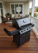 A BBQ Boards® Napoleon Prestige 500 Side Cutting Board by BBQ Boards® offers extra grill prep space on a black outdoor gas grill with a wooden side shelf, set on a porch with white railings and nearby seating area.