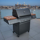 A Napoleon Prestige 500 gas grill with black finish features BBQ Boards® Connected Side Cutting Boards (Matching Pair) on a rooftop balcony, city skyline visible in the twilight background.